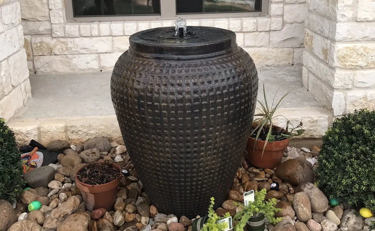 Bronze-colored ceramic waffle fountain with water flowing over the sides into a pile of rocks with a stone house in the background