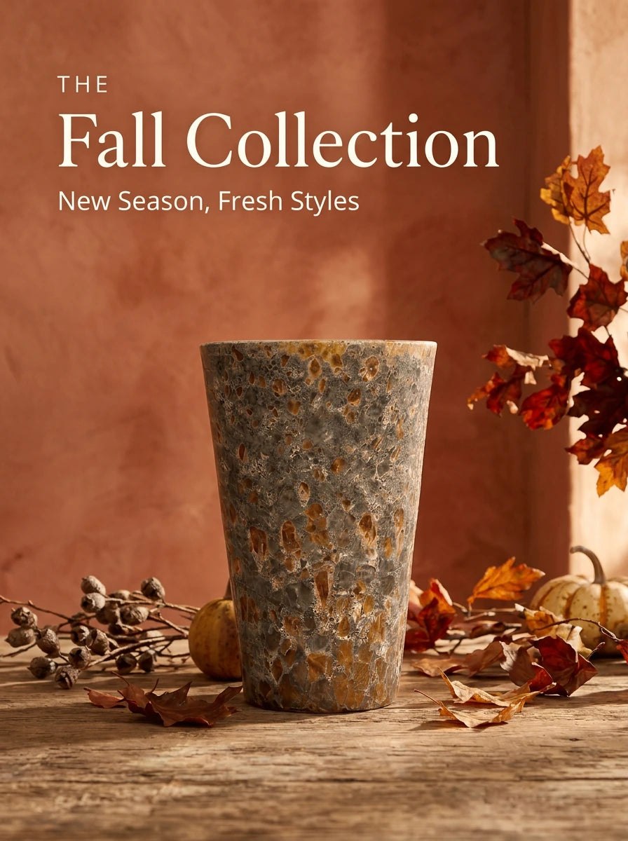 A tall, tapered ceramic planter with a speckled gray-and-orange glaze, surrounded by autumn leaves, dried seed pods, and small pumpkins, set against a warm terracotta wall.