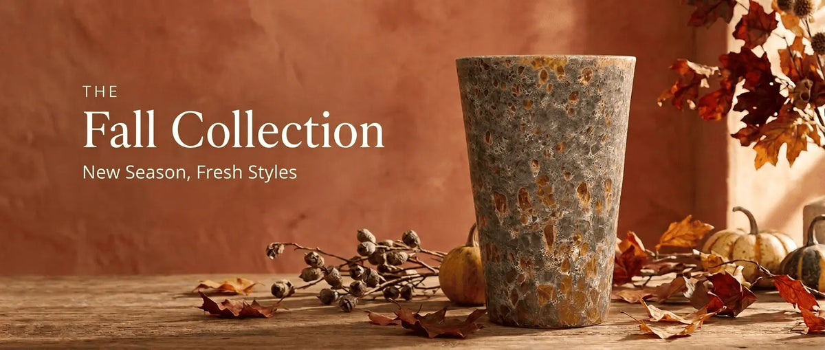 A tall, tapered ceramic planter with a speckled gray-and-orange glaze, surrounded by autumn leaves, dried seed pods, and small pumpkins, set against a warm terracotta wall.