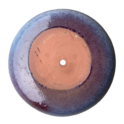 Underside of a falling purple ceramic succulent bowl, showing an unglazed terracotta base with a central drainage hole, ringed by plum glaze that fades to speckled periwinkle at the edge